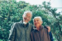 A smiling, elderly asian man with glasses with his arm around his smiling wife.