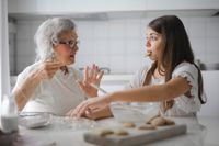 An older lady making cookies with young girl that has a cookie in her mouth.